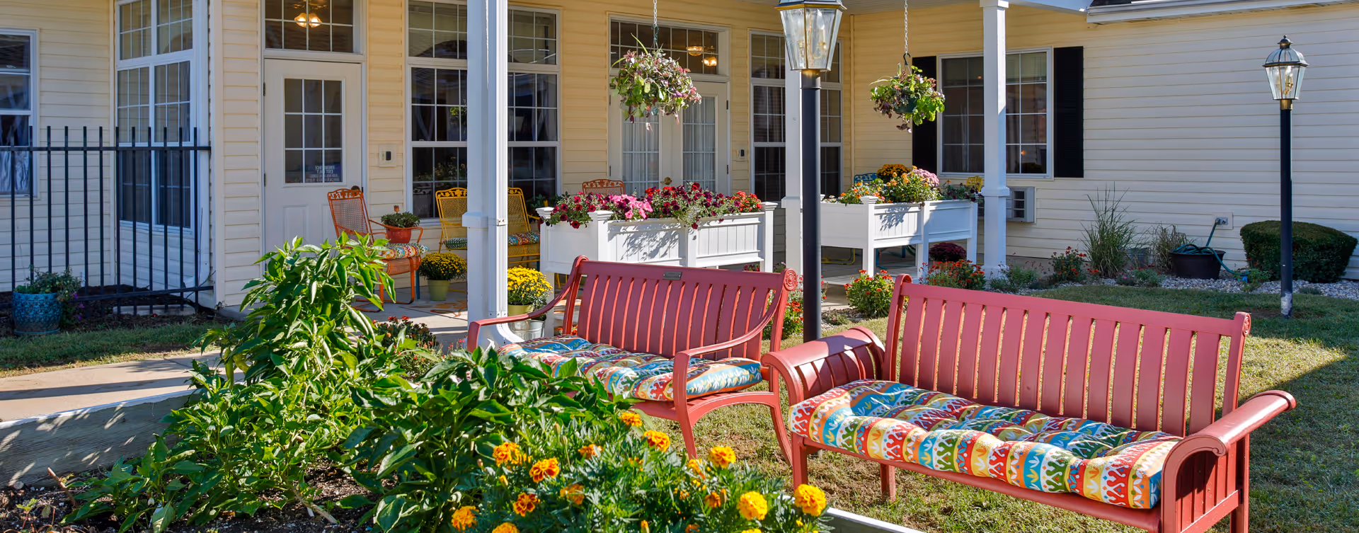 Outdoor seating area at a senior living facility with two red benches featuring colorful cushions, surrounded by flower beds with yellow and pink flowers. The building exterior is light yellow with multiple windows and a door leading inside. Hanging flower baskets and lamp posts are also visible.