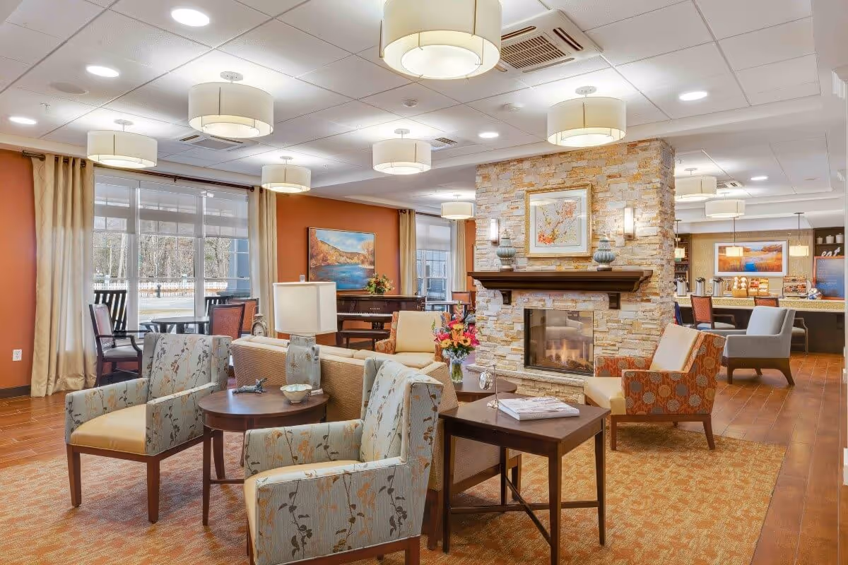 A spacious and well-lit living room area in a senior living facility featuring multiple upholstered chairs and sofas arranged around a stone fireplace with a wooden mantle. The room has large windows with beige curtains, warm-toned walls, and ceiling lights. There is a table with a lamp and flowers, and a dining area with chairs and tables is visible in the background.