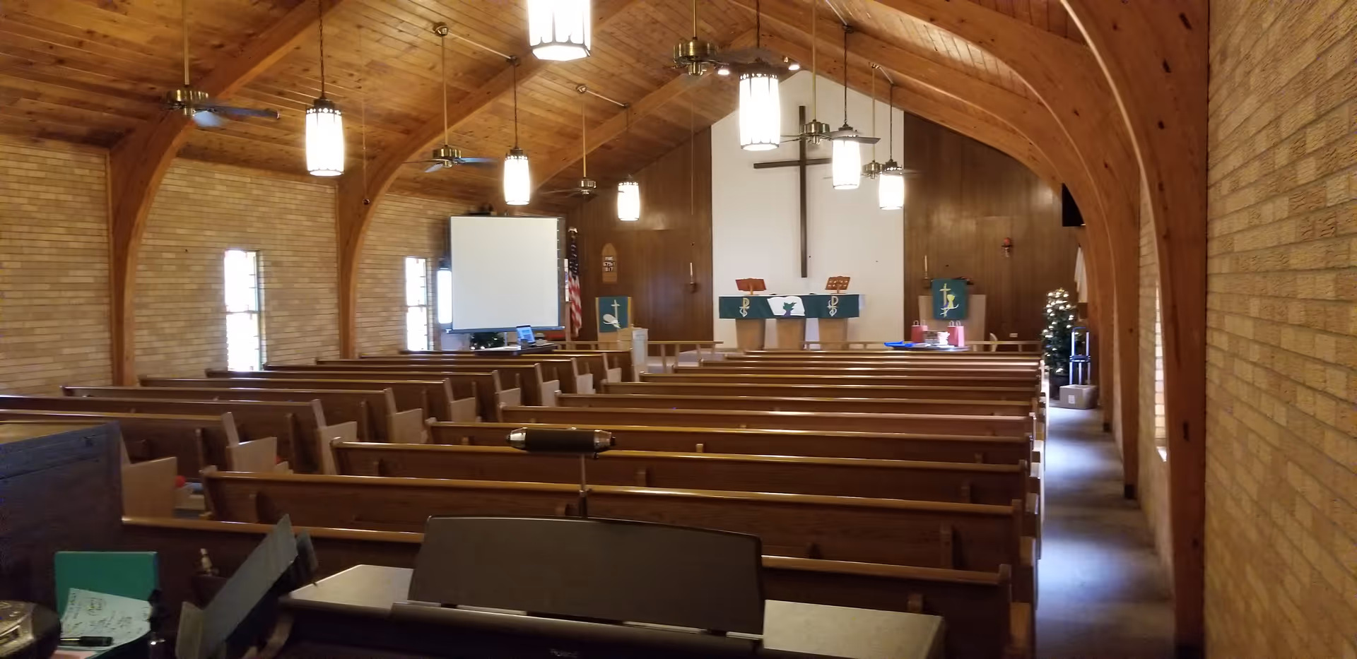 Interior view of a chapel with wooden pews, arched wood ceiling, pendant lights, and an altar with a cross.