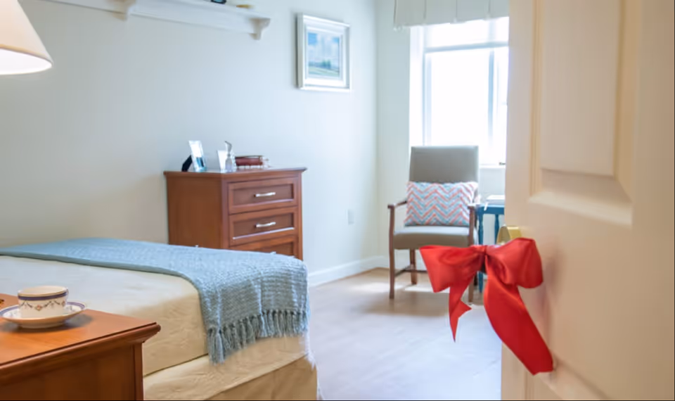 A bright and cozy bedroom in a senior living facility featuring a neatly made bed with a light blue throw blanket, a wooden nightstand with a cup and saucer, a wooden dresser with framed photos and books, and a comfortable armchair with a decorative pillow near a window. A red ribbon is tied to the door handle.