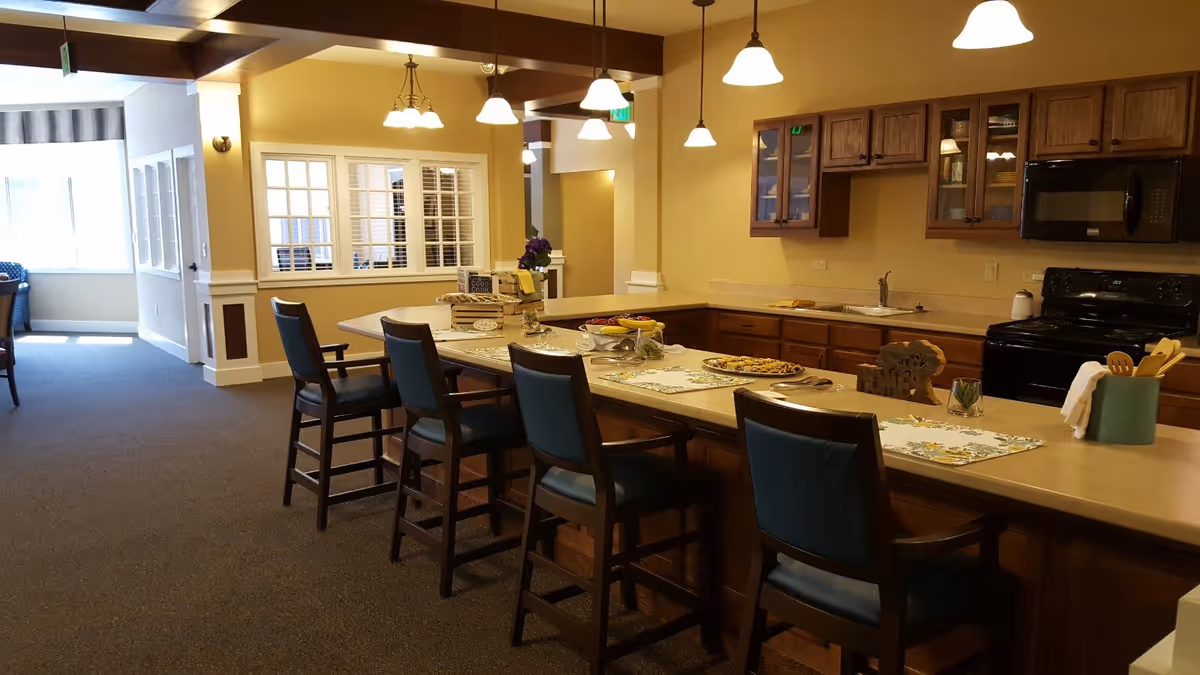 Open communal kitchen and dining area with a long island, several bar stools, pendant lights, and wooden cabinetry.