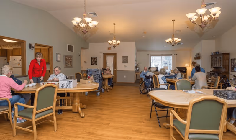 A communal dining area in a senior living facility with several elderly residents seated at tables, some in wheelchairs, engaging in activities and socializing. A staff member in a red sweater is assisting one of the residents. The room has wooden floors, multiple chandeliers, and a window letting in natural light.