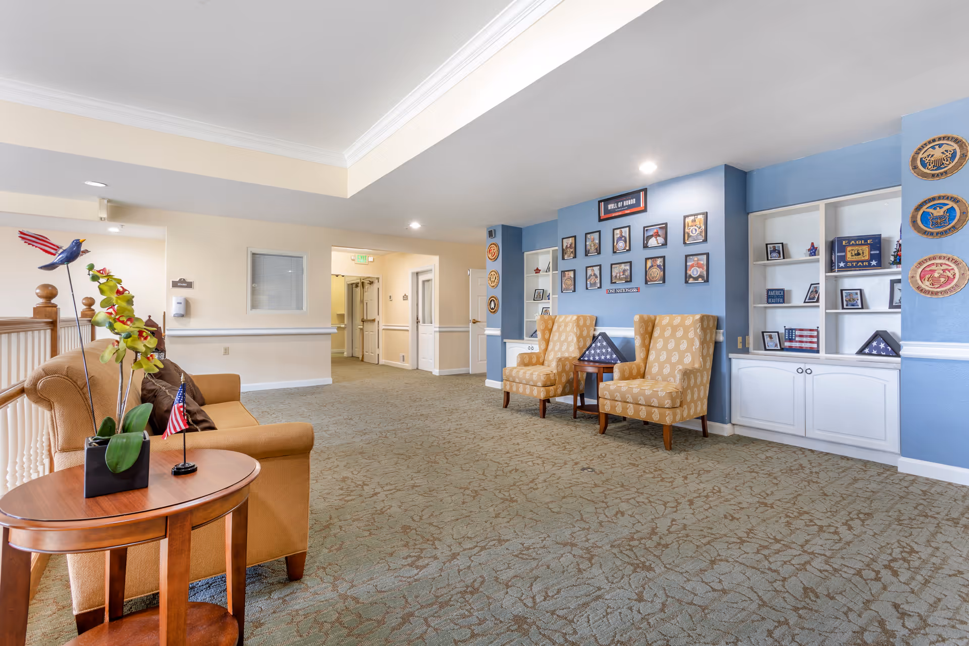A spacious interior hallway area in a senior living facility with beige carpet and light-colored walls. There is a brown sofa with a small wooden side table holding a potted orchid and a small American flag. Two patterned armchairs flank a small table displaying a folded American flag. The blue accent wall behind the chairs features a 'Wall of Honor' with framed photos and military insignias. Built-in white shelves display various patriotic decorations and memorabilia.