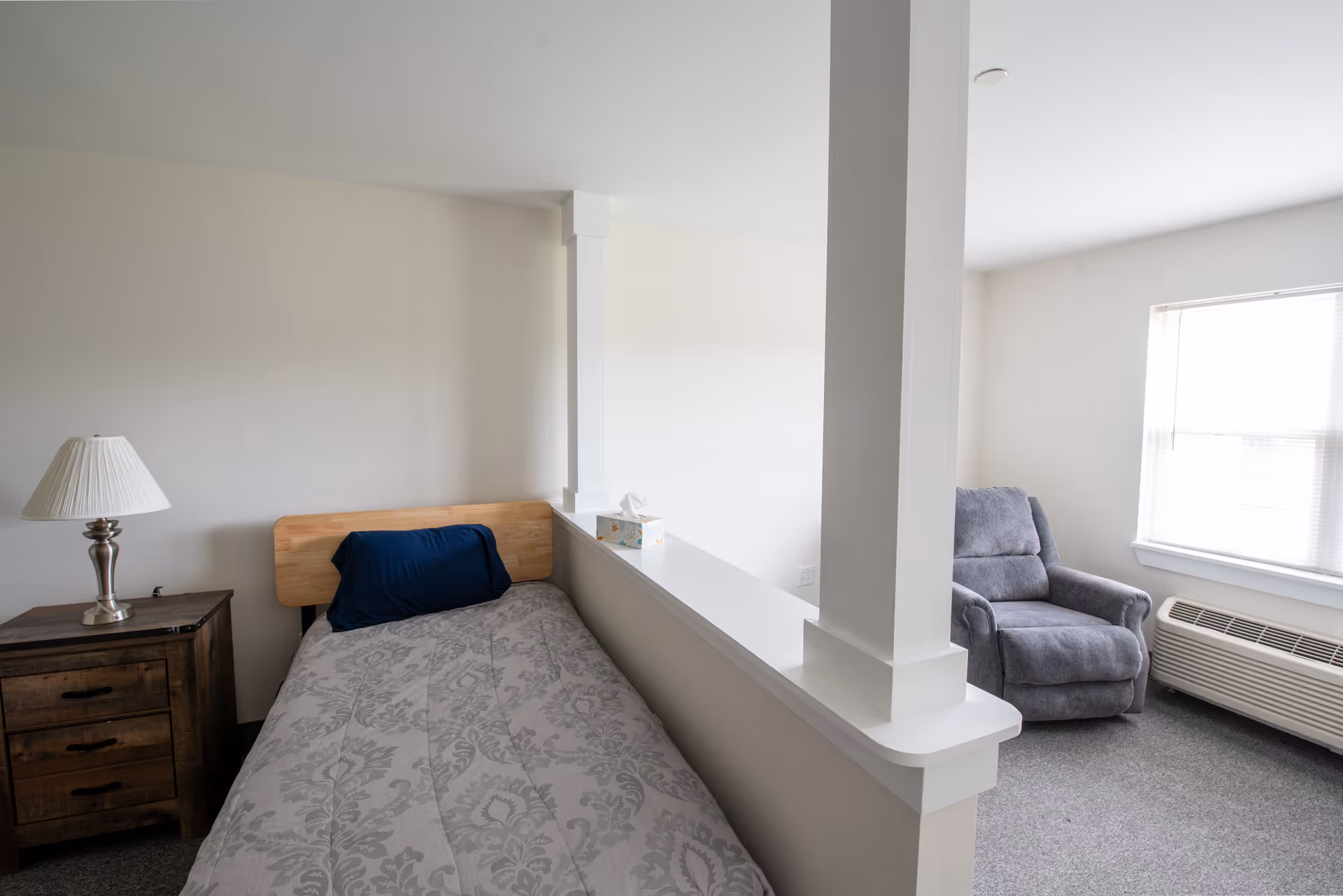 Small bright bedroom with a single bed and nightstand on one side of a half wall and a gray recliner beside a window on the other.