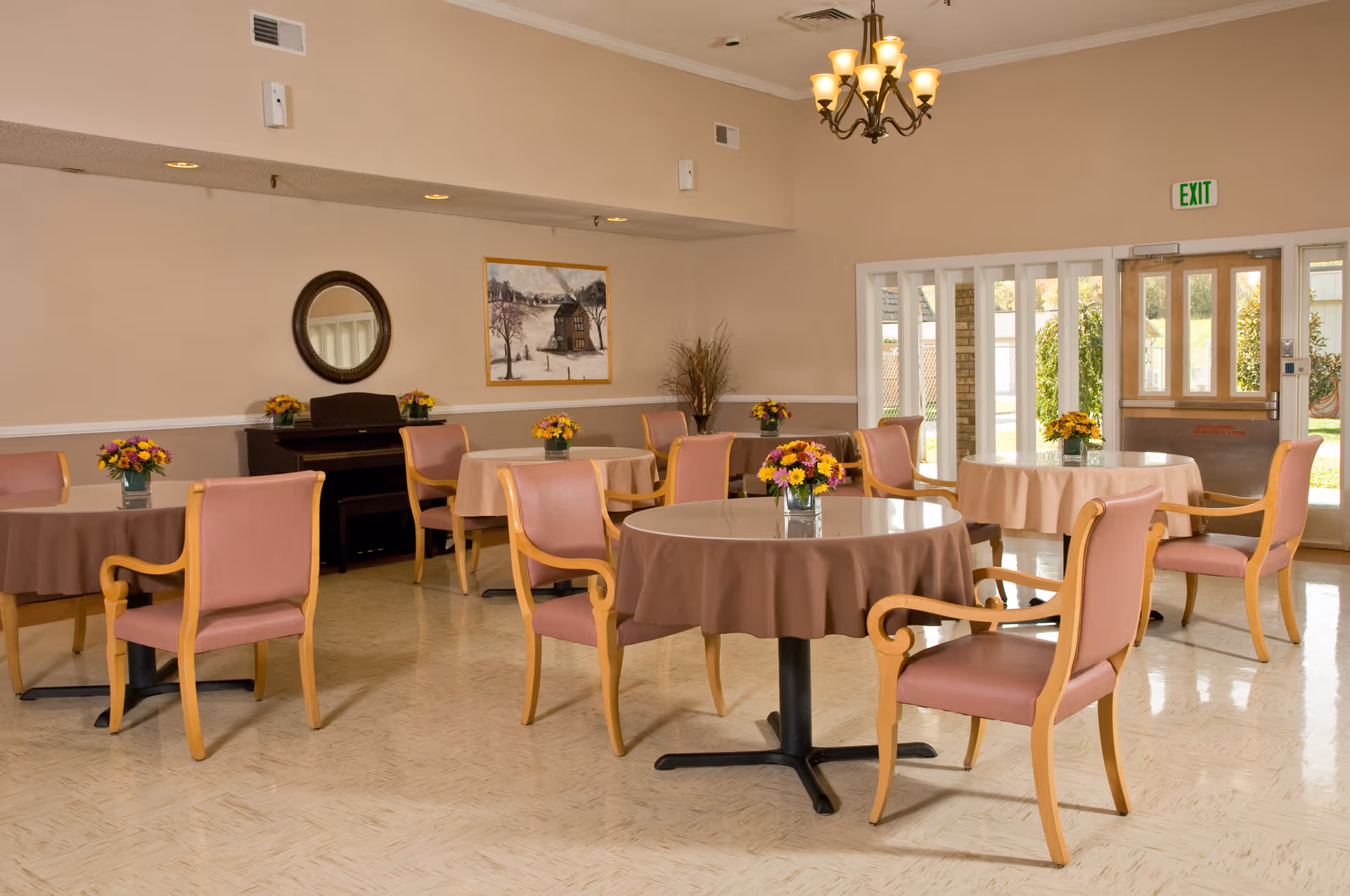 Bright communal dining room with round tables, pink upholstered chairs, floral centerpieces and an exit door.