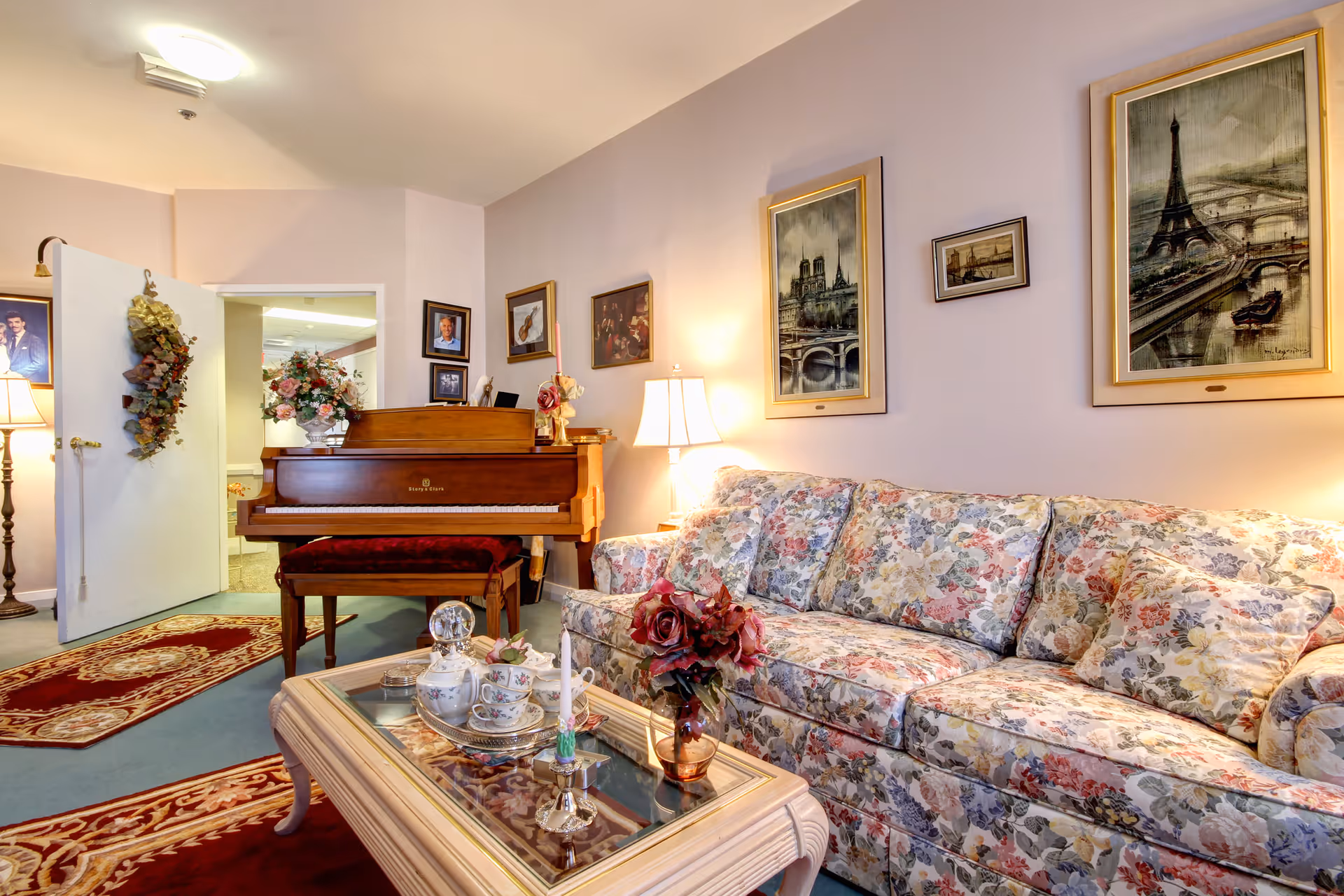 A cozy living room with a floral patterned sofa and matching cushions, a glass-top coffee table with a tea set and a vase of flowers, a wooden piano with a red cushioned bench, framed paintings on the walls, and a standing lamp providing warm light. The room has a soft pastel wall color and decorative rugs on the floor.