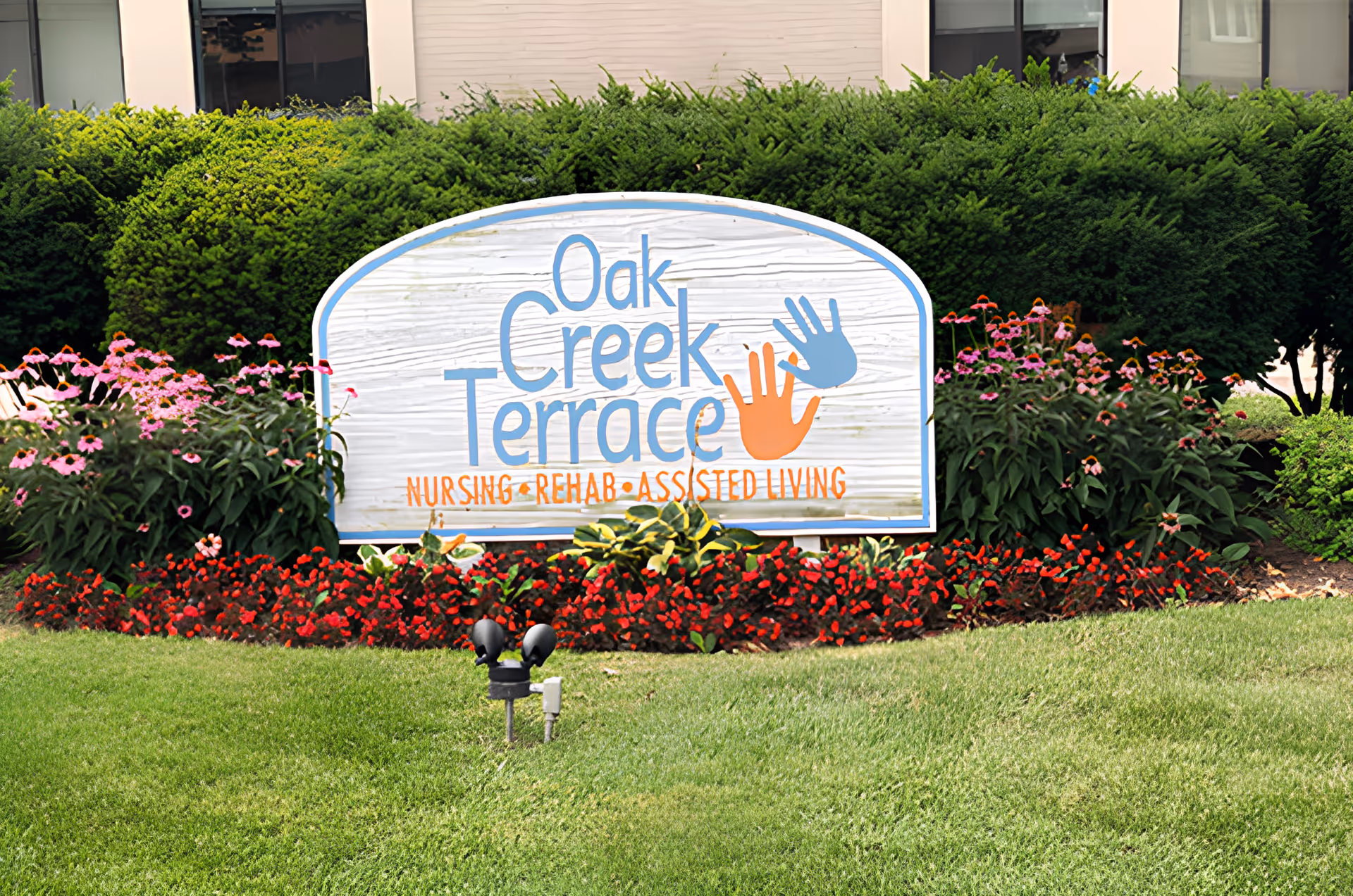A painted wooden sign for Oak Creek Terrace Nursing • Rehab • Assisted Living surrounded by flowers and shrubs on a lawn in front of the building.
