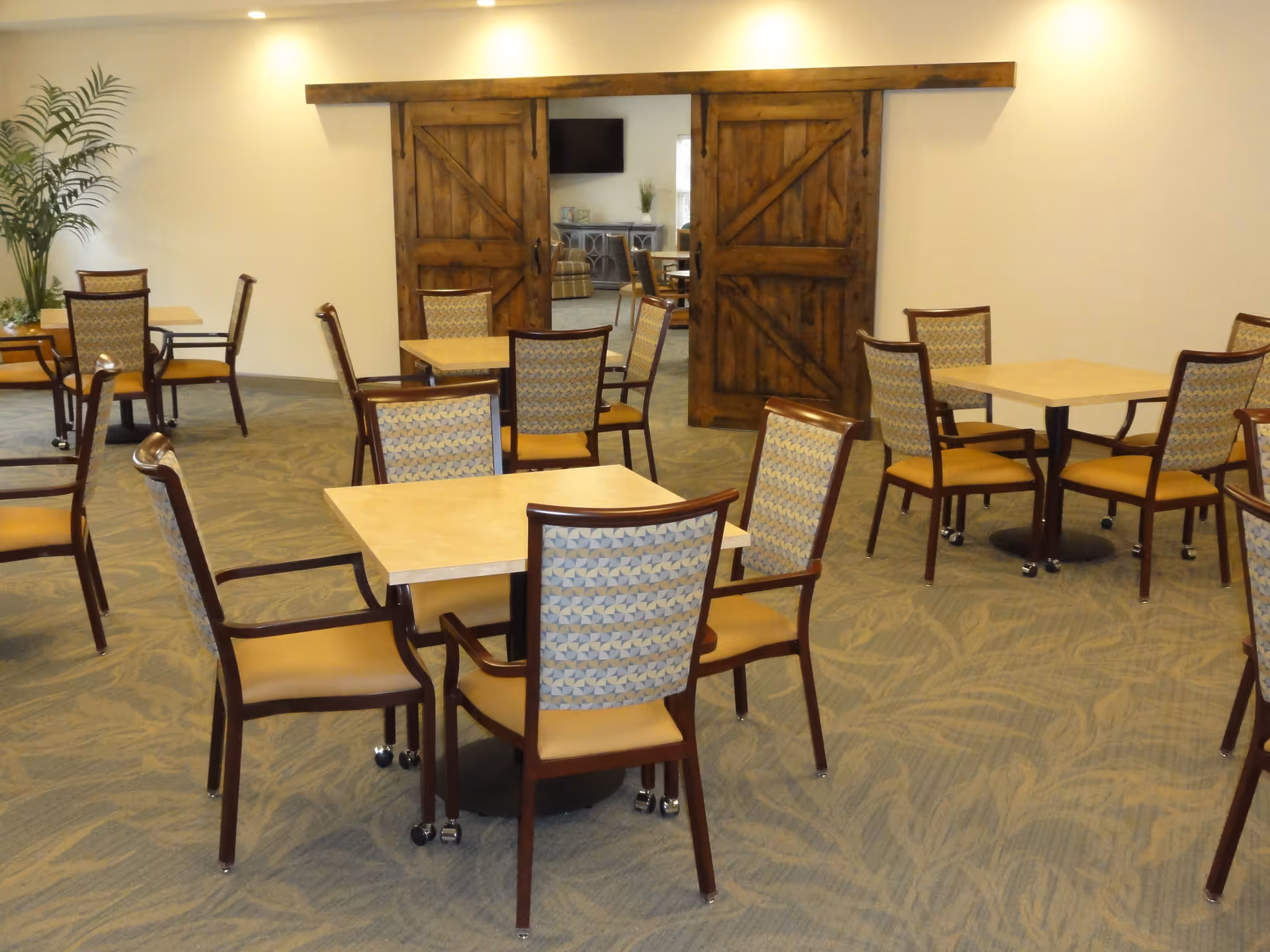 A dining area with several square tables and cushioned chairs arranged neatly on a carpeted floor. In the background, there are two large wooden sliding barn doors partially open, revealing a lounge area with a TV and additional seating. A tall green plant is placed in the corner of the room.