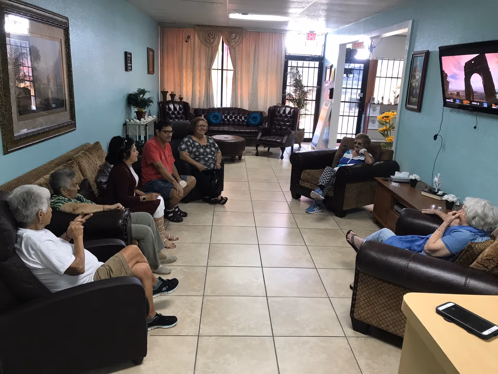 A group of elderly people and one younger man sitting in a living room area of a senior daycare facility. The room has light blue walls, tiled floors, several armchairs and sofas arranged along the walls, a TV mounted on the wall, and framed pictures. There is a window with curtains and a door at the far end of the room.