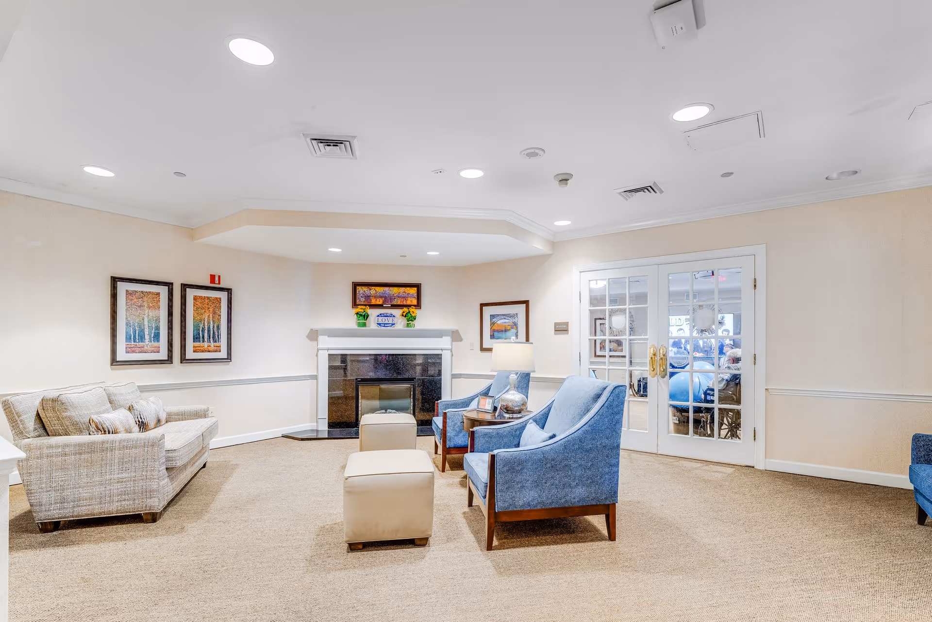 A cozy living room area in a senior living facility featuring a beige sofa, two blue armchairs, two beige ottomans, a fireplace with decorative items on the mantel, framed artwork on the walls, and glass double doors leading to another room with exercise balls visible.