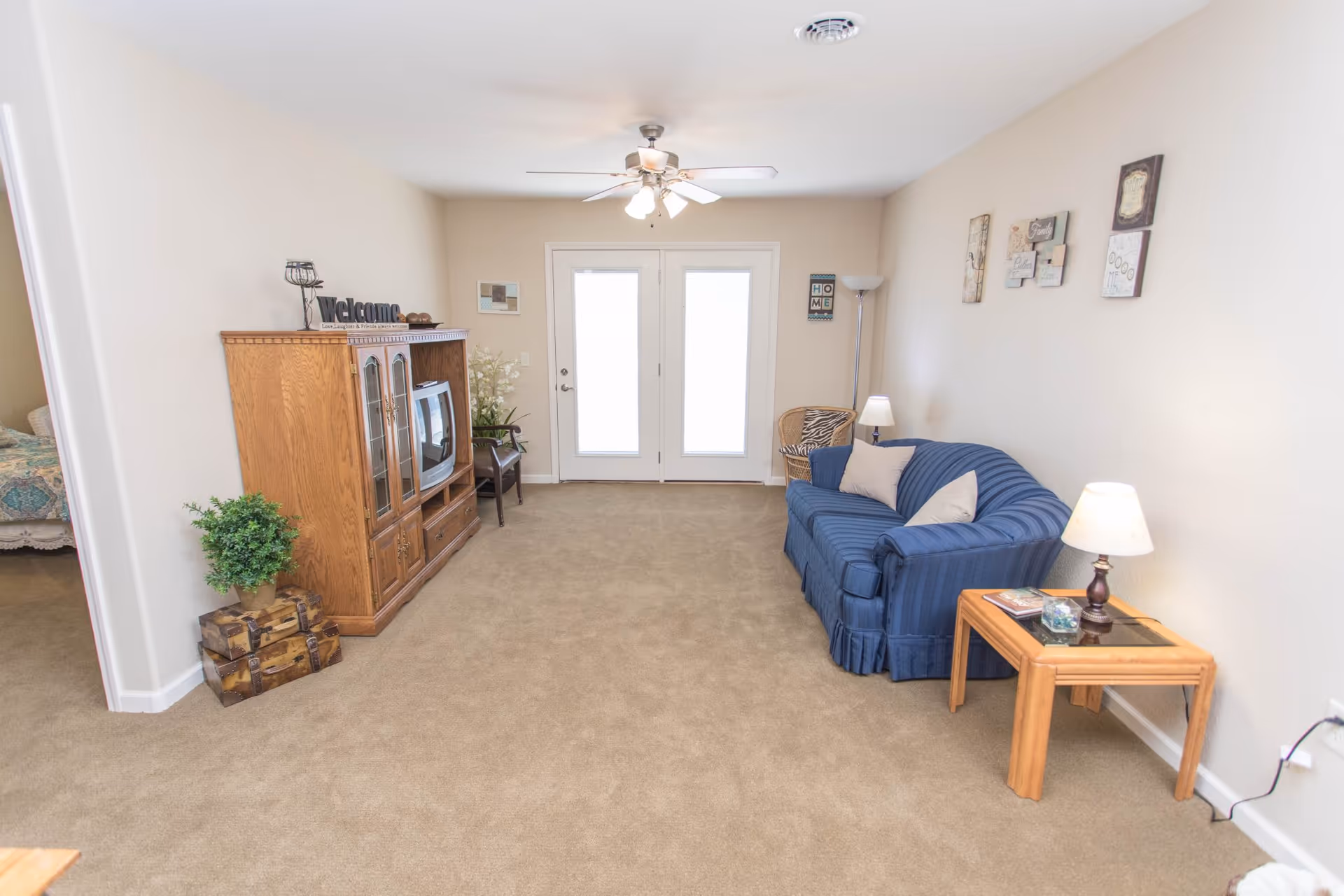 Living room with a blue couch, wooden TV cabinet, side table with lamp, and double glass doors at the far wall.