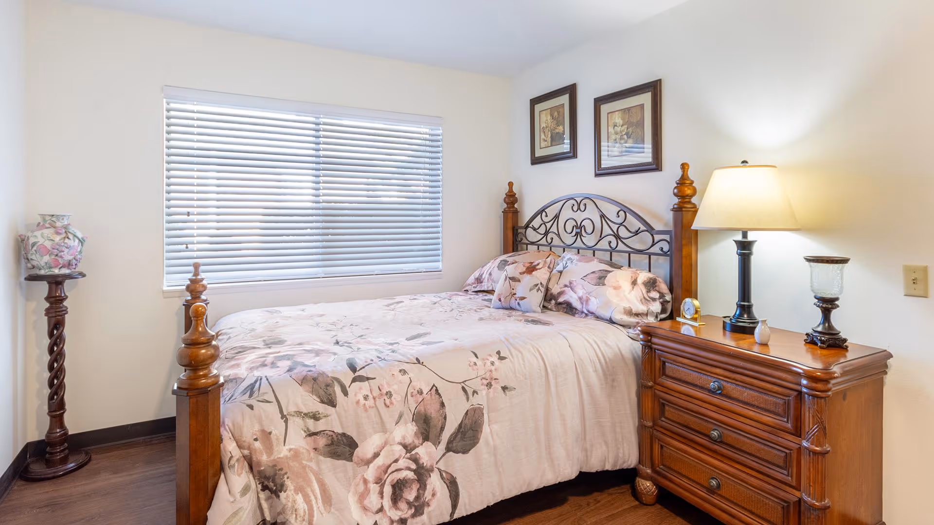 A cozy bedroom with a wooden bed frame featuring an ornate metal headboard. The bed is made with floral-patterned bedding and matching pillows. Next to the bed is a wooden nightstand with three drawers, a table lamp, a small clock, and decorative items. On the left side of the room, there is a tall wooden pedestal holding a floral vase. A window with closed blinds is behind the bed, and two framed floral artworks hang on the wall above the headboard.
