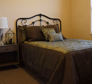 A neatly made bed with a decorative metal headboard, covered with a green and brown patterned bedspread and several pillows. Next to the bed is a wooden nightstand with a white lamp and a decorative glass terrarium. The room has beige walls and a window with blinds partially visible.