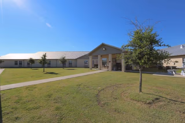 Exterior view of Stillhouse Rehabilitation And Healthcare Center showing a single-story building with a metal roof, a covered entrance with seating, a paved walkway, and a grassy lawn with several small trees under a clear blue sky.