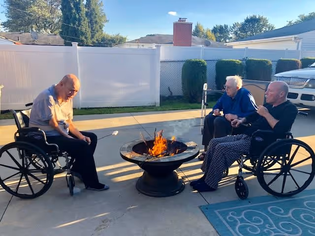 Three elderly men in wheelchairs sitting around a fire pit outdoors, roasting marshmallows on sticks. They are in a paved backyard area with a white fence, some bushes, and parked cars in the background under a clear sky.