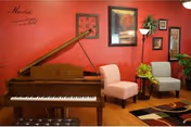 Interior room with a wooden grand piano against a red wall, two upholstered chairs in pink and gray, framed artwork on the wall, a small table with a plant, and a colorful rug on a wooden floor.
