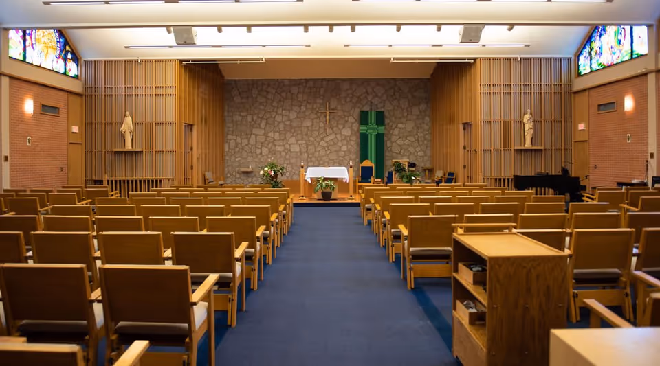 Interior view of a chapel with rows of wooden chairs facing an altar. The altar is adorned with flowers and a white cloth, and a cross is mounted on a stone wall behind it. There are statues on either side of the altar and stained glass windows near the ceiling.