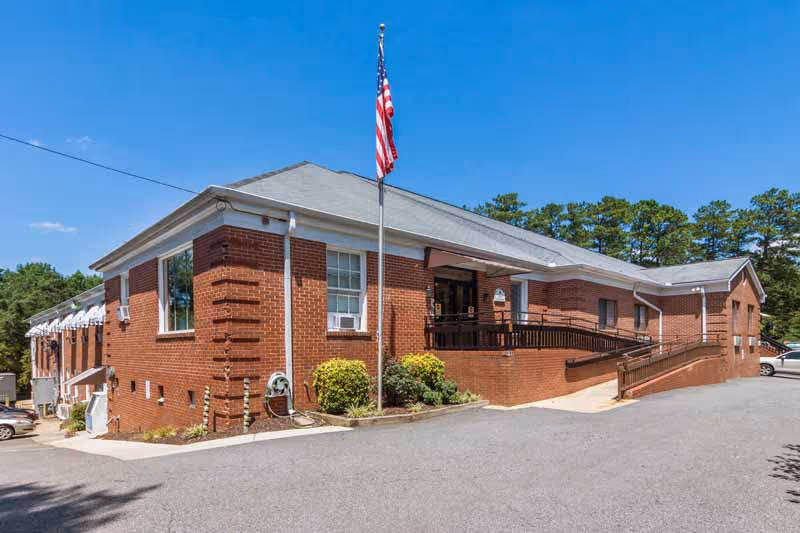 Exterior view of a single-story brick building with a gray roof, an American flag on a flagpole in front, a wheelchair ramp leading to the entrance, and some shrubs along the building. The sky is clear and blue.