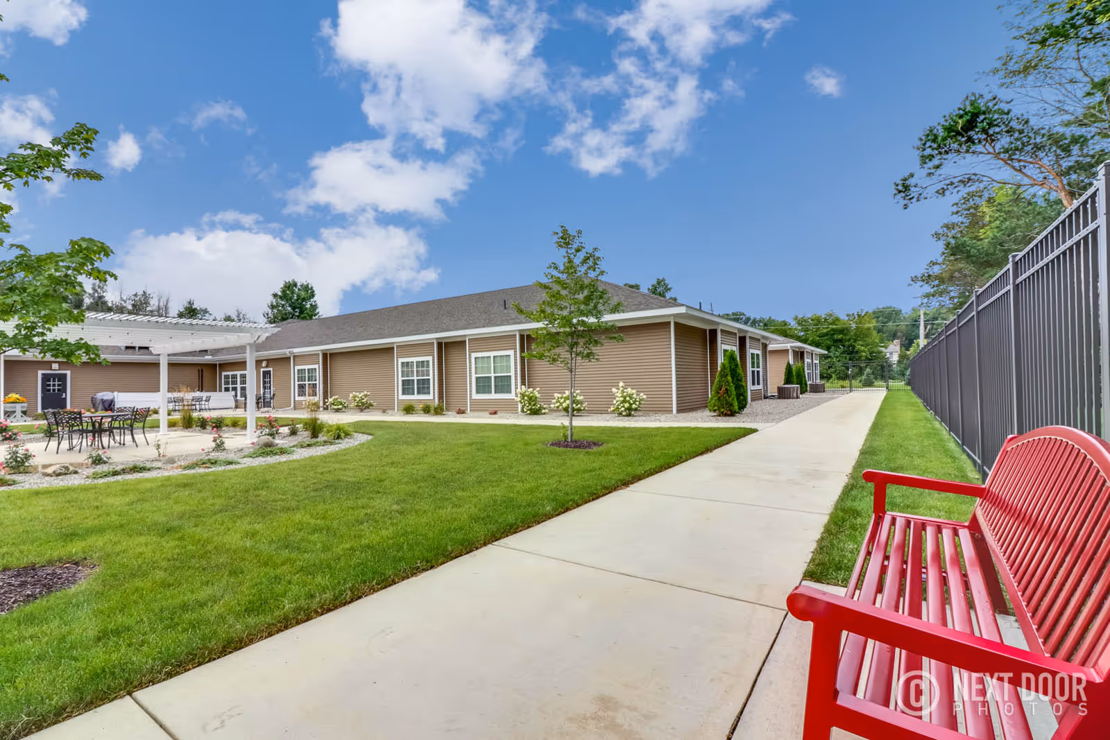 Outdoor view of a single-story assisted living facility building with beige siding and white trim. There is a concrete walkway leading alongside the building, a red bench on the right side, and a patio area with tables and chairs under a white pergola on the left. The area is landscaped with green grass, small trees, and flowering plants under a partly cloudy blue sky.