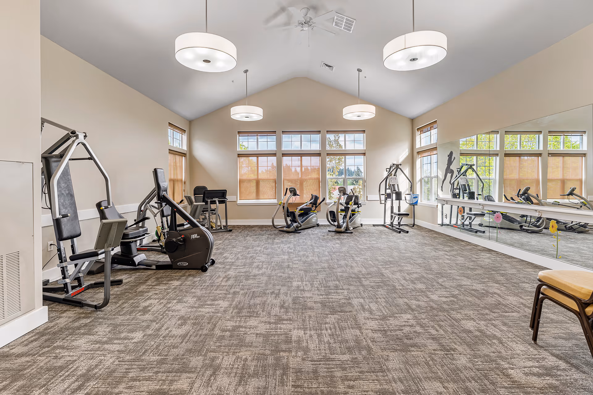 Bright fitness room in a senior living facility with exercise machines, a mirrored wall, and large windows.