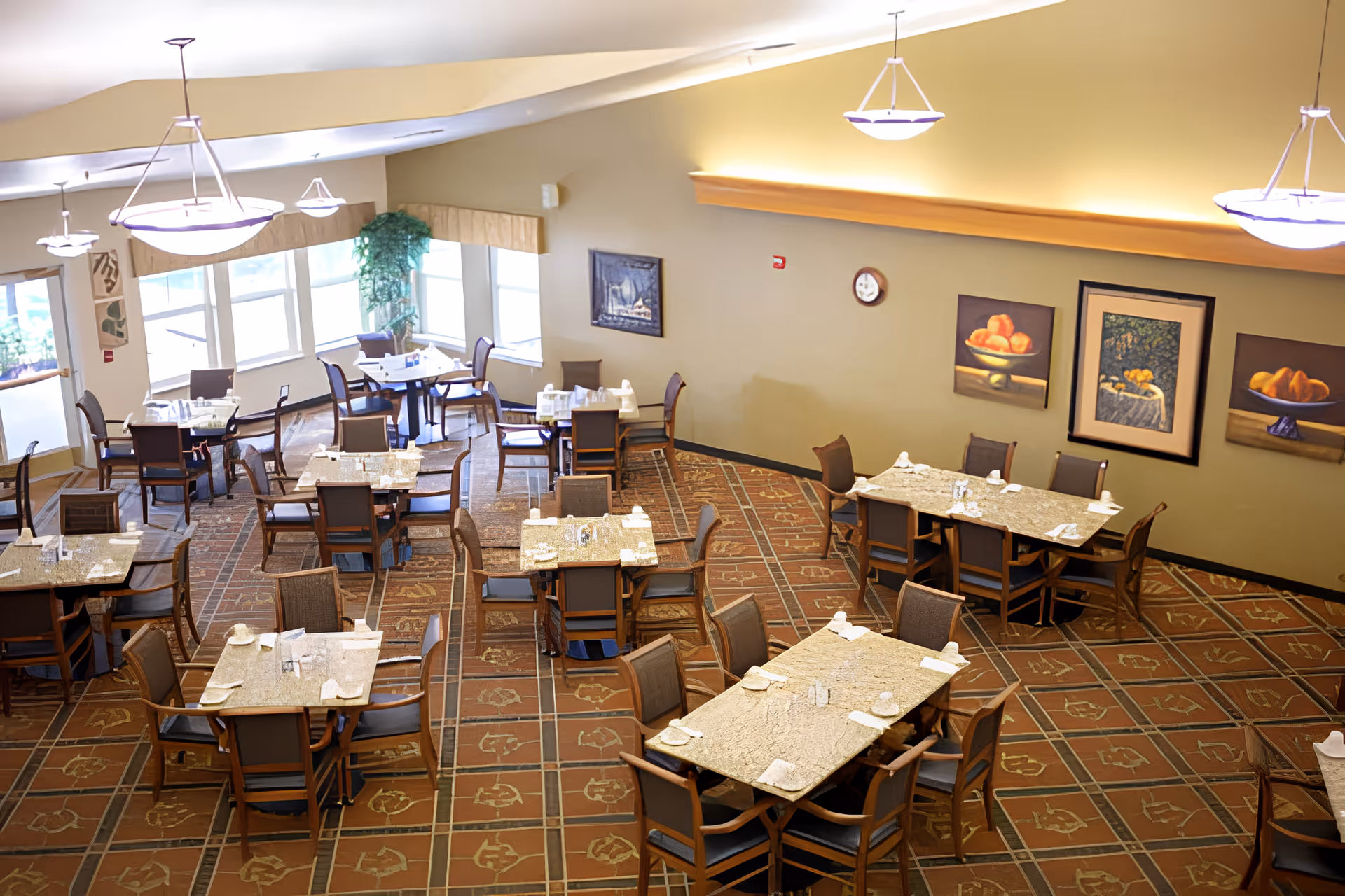 A spacious dining room with multiple tables and chairs arranged neatly. The tables are set with napkins and utensils. The room has large windows allowing natural light to enter, decorative paintings on the walls, and hanging ceiling lights providing illumination.