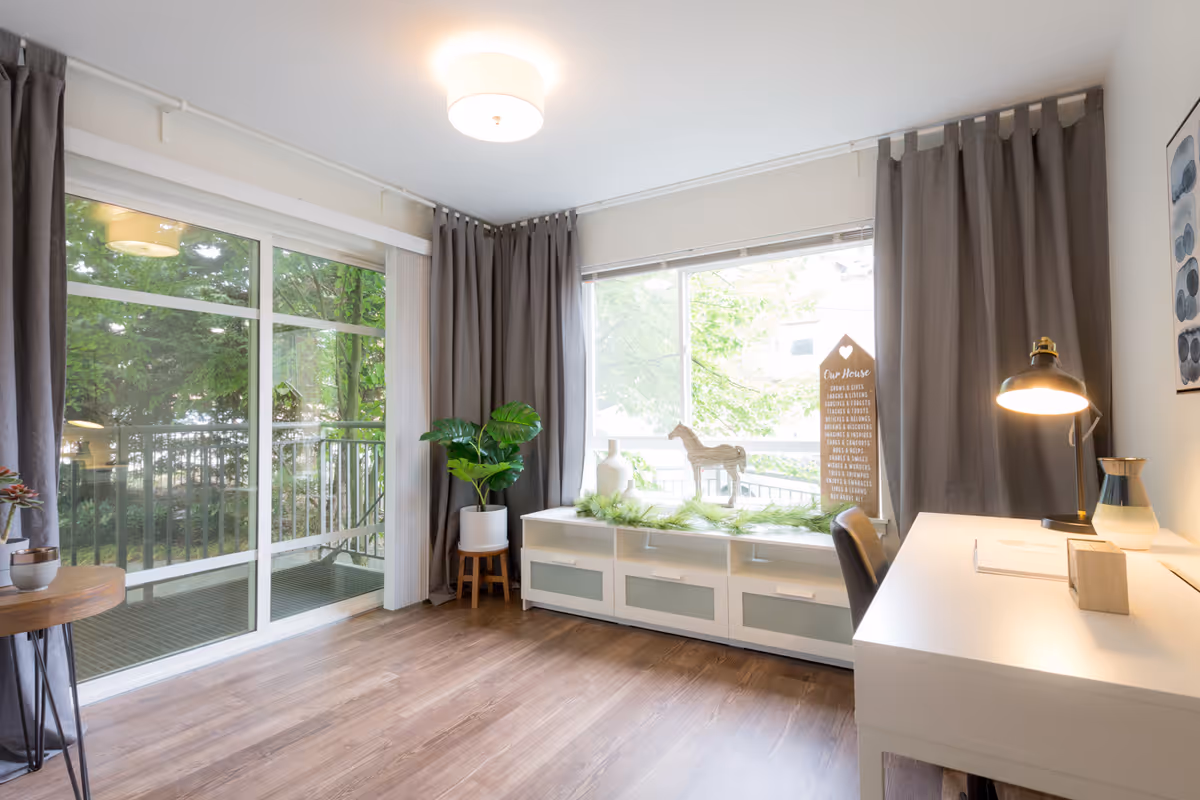 A bright room with large windows covered by gray curtains, a white desk with a lit desk lamp, a chair, a white cabinet with decorative items including a small horse figurine and a sign, and a potted plant on a wooden stand. Outside the windows, green trees and a balcony railing are visible.
