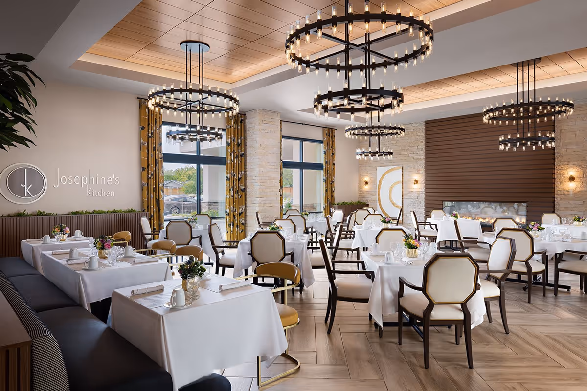 A bright and elegant dining room with multiple tables covered in white tablecloths, set with cups, glasses, and floral centerpieces. The room features large windows with yellow patterned curtains, modern circular chandeliers hanging from a recessed ceiling, and a fireplace with a wooden accent wall. A sign on the wall reads 'Josephine's Kitchen'.