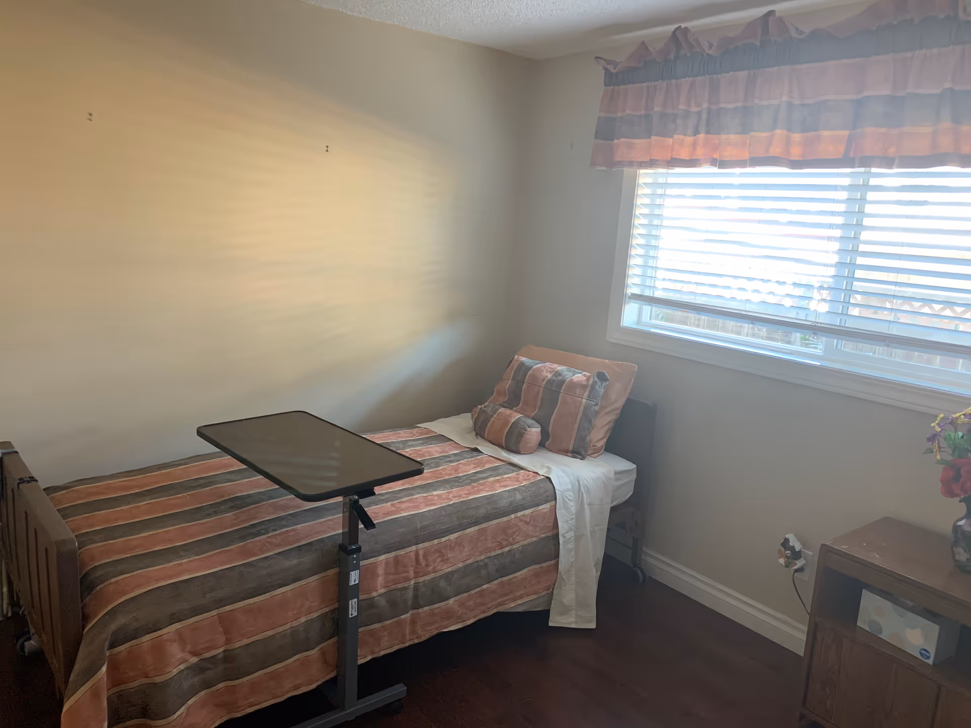 A small bedroom with a single bed covered in a striped bedspread in shades of brown, orange, and beige. The bed has matching pillows and a white sheet partially draped over it. Next to the bed is a small adjustable overbed table. A window with blinds and a valance lets in natural light. There is a wooden nightstand with a vase of flowers and a box of tissues on it. The walls are plain and light-colored, and the floor is dark wood.