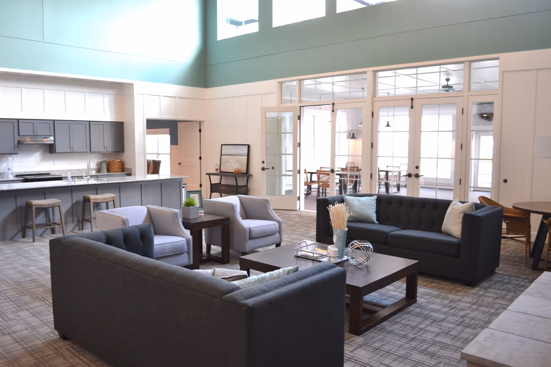 A spacious and well-lit living area featuring two dark gray sofas, two light gray armchairs, a dark wooden coffee table with decorative items, and a kitchen area with gray cabinets and a granite countertop with three bar stools. Large windows and glass doors lead to another room with tables and chairs.