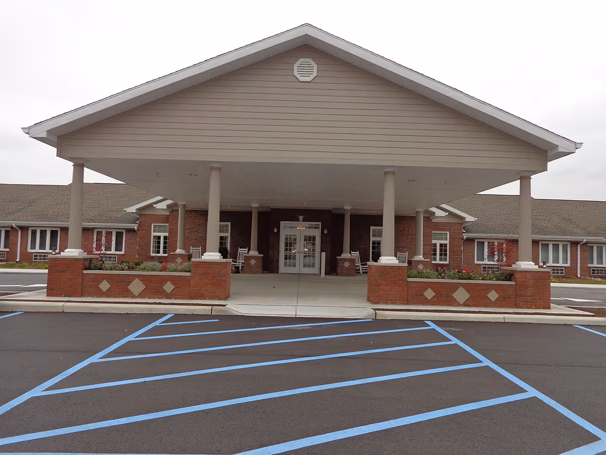 Front entrance of a senior living building with a large covered porte-cochere, brick columns and marked accessible parking spaces.
