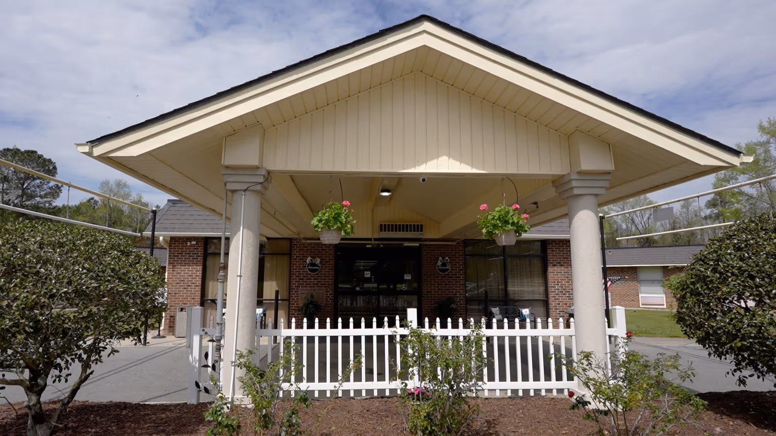 Front entrance of Mary Gran Nursing Center showing a covered porch with two columns, hanging flower pots with pink flowers, a white picket fence, and brick exterior walls under a partly cloudy sky.