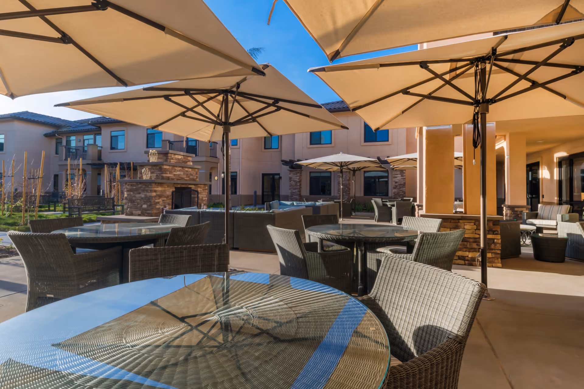 Outdoor patio with round glass-topped tables, wicker chairs, and large umbrellas in front of a multi-story senior living building.