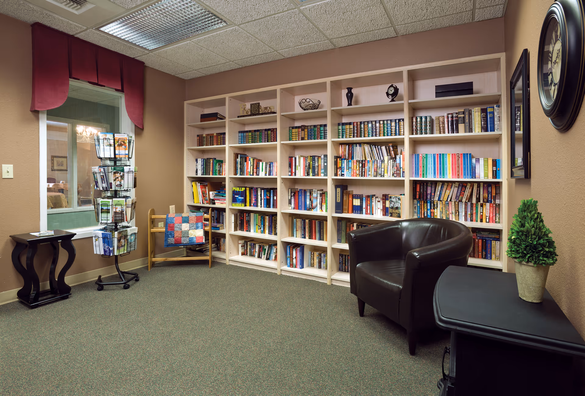 A cozy reading room with a large bookshelf filled with books, a black leather armchair, a small black table with a potted plant, a wooden chair with a colorful quilt draped over it, and a rotating rack with brochures near a window with a red valance.
