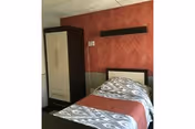 A small bedroom with a single bed covered with a patterned blanket and a reddish throw. There is a wardrobe with two doors next to the bed, and the wall behind the bed has a textured reddish wallpaper with a black horizontal shelf mounted above the headboard.