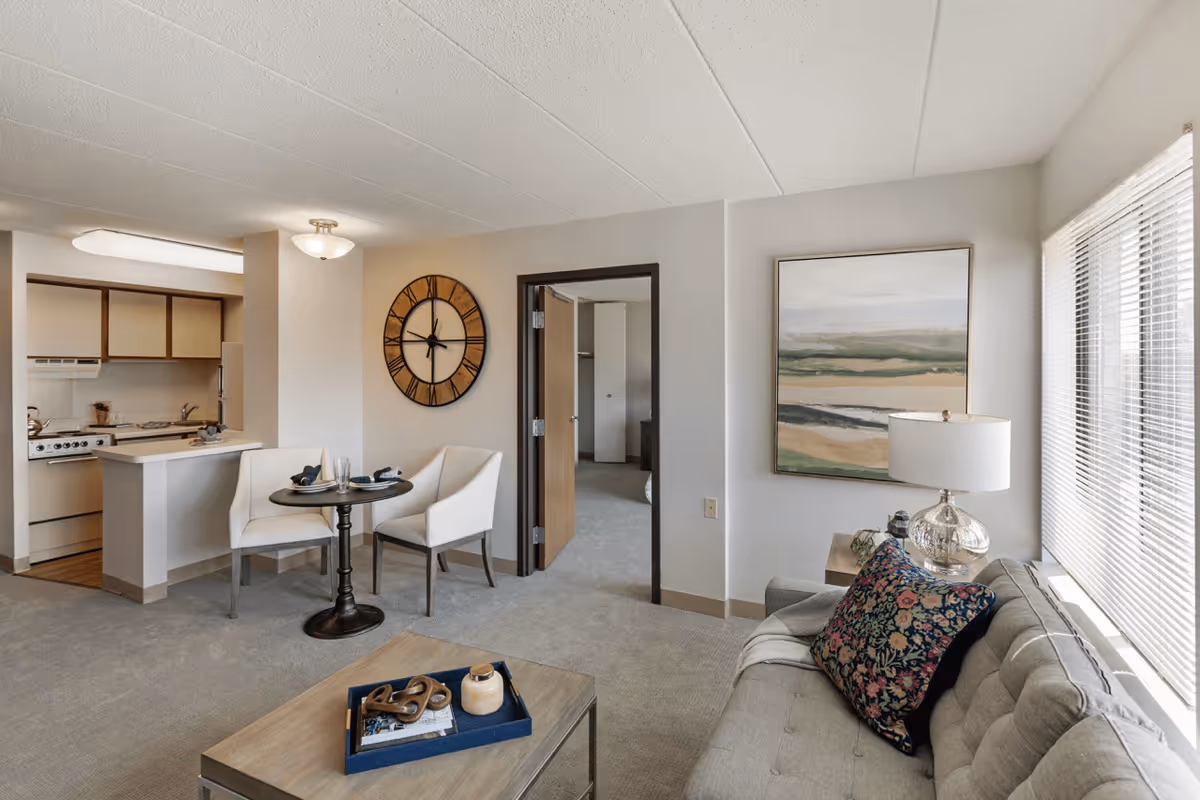 A cozy senior living apartment interior featuring a small round dining table with two white chairs, a wall clock, a kitchen area with stove and cabinets, a living room with a gray sofa adorned with a floral pillow, a wooden coffee table with decorative items, a side table with a lamp, and a large window with blinds letting in natural light.