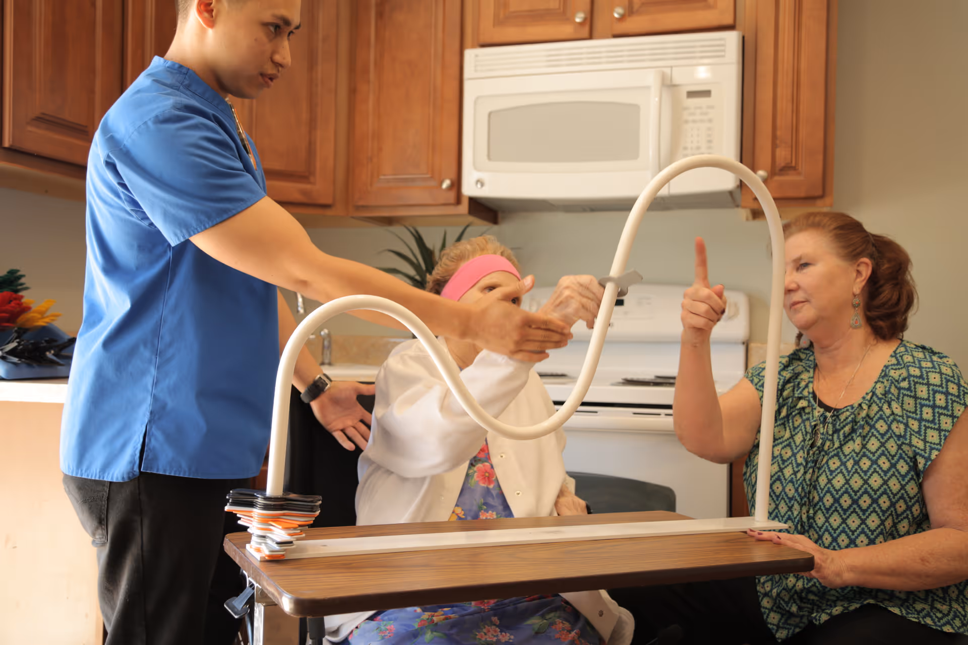 A young male caregiver in a blue uniform assists an elderly woman in a wheelchair as she interacts with a middle-aged woman using a white curved therapy device in a kitchen setting with wooden cabinets and a microwave in the background.