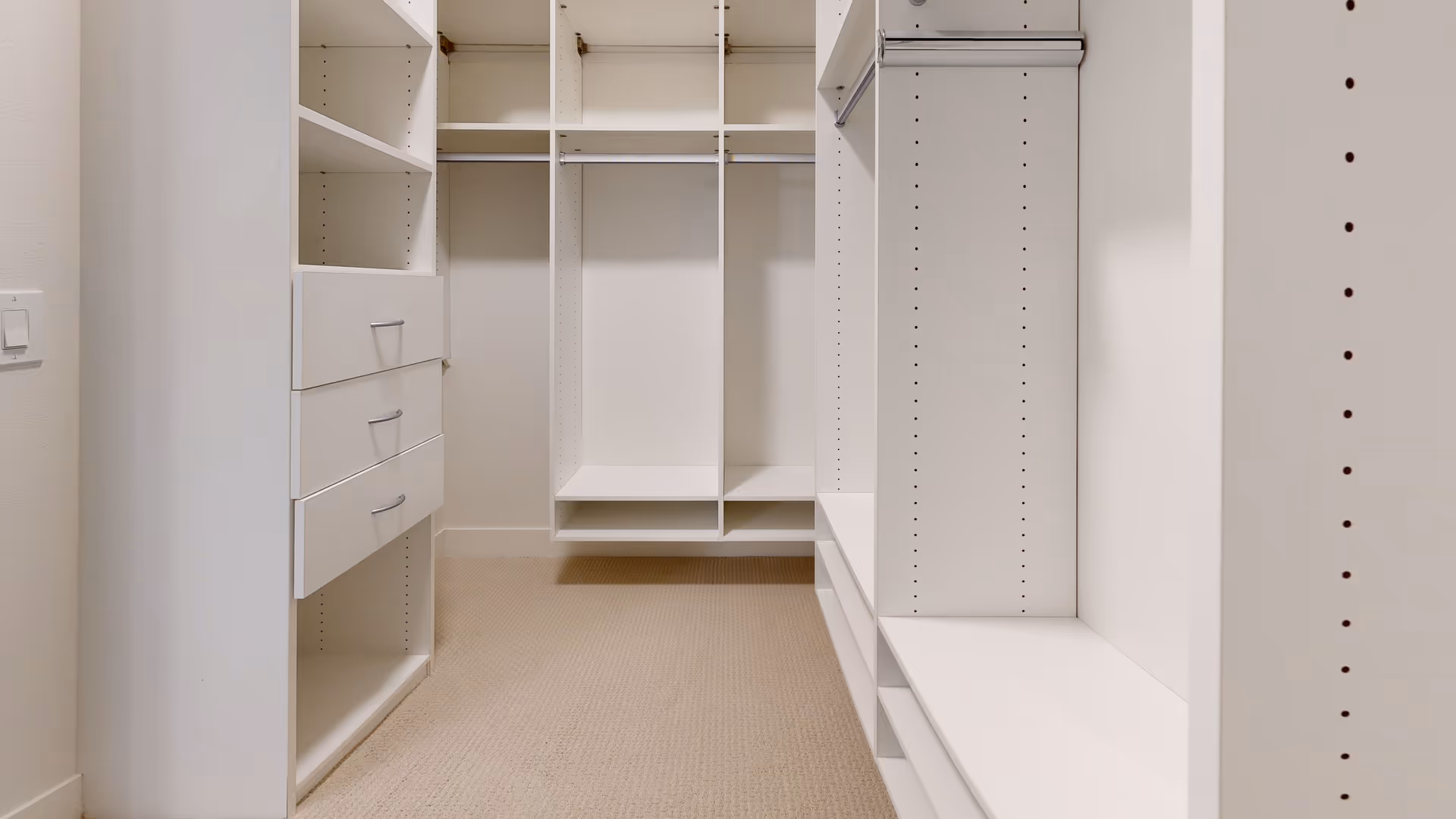 Empty walk-in closet with white built-in shelves, drawers, and hanging rods, beige carpeted floor, and white walls.