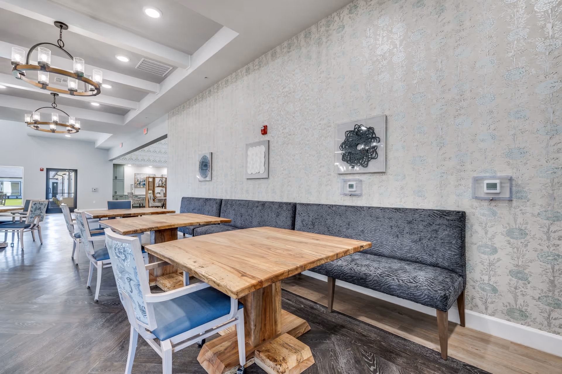 Interior view of a dining area in The Lodge of Saginaw Health & Wellness featuring wooden tables with blue cushioned chairs and a long upholstered bench against a patterned wall. The room has decorative light fixtures hanging from the ceiling and artwork on the walls.