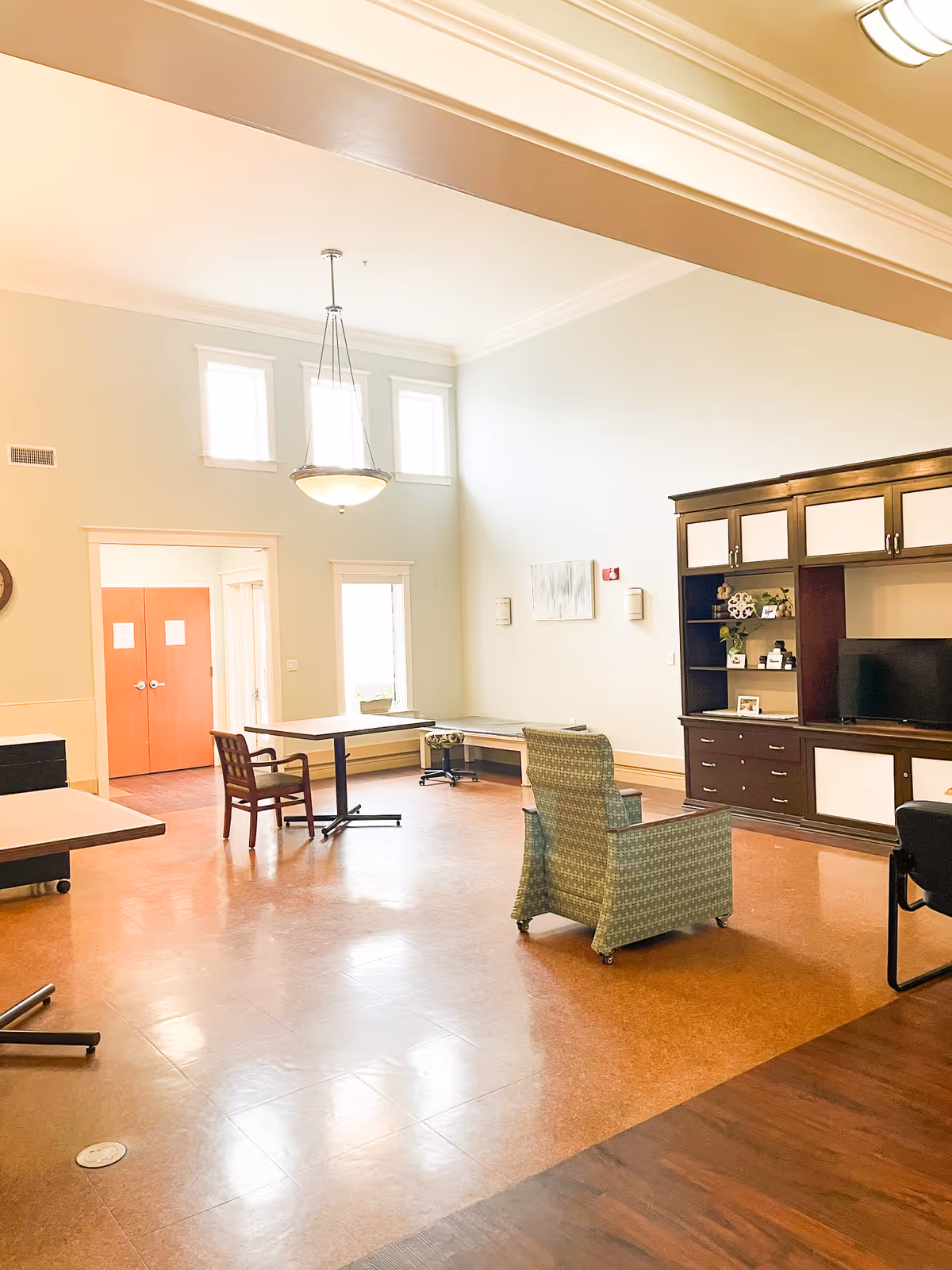 A spacious, well-lit common area with high ceilings and large windows. The room features a green patterned armchair, wooden chairs, tables, a built-in dark wood entertainment center with a TV, and light-colored walls. There is a hanging light fixture and double doors leading to another room.