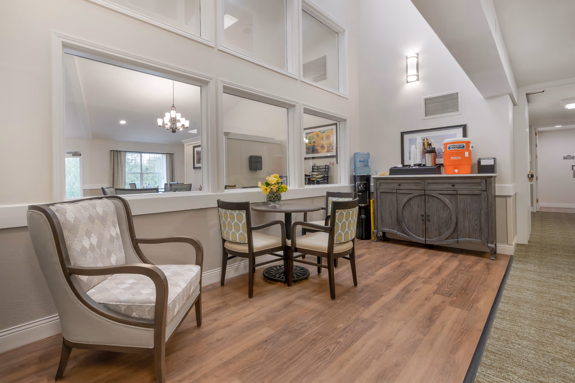 A bright common seating area with upholstered chairs around a small round table, a wooden sideboard, and a water cooler in a hallway.