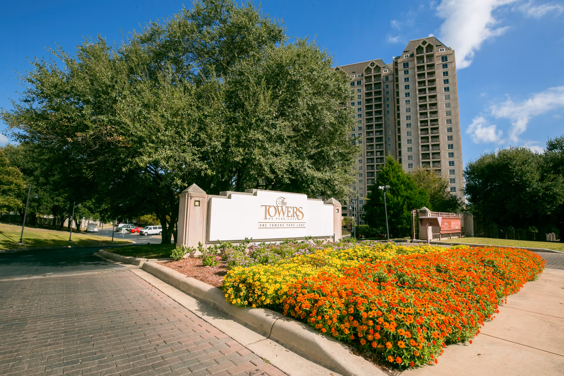 Entrance sign and colorful flowerbeds in front of a tall residential tower building under a blue sky.
