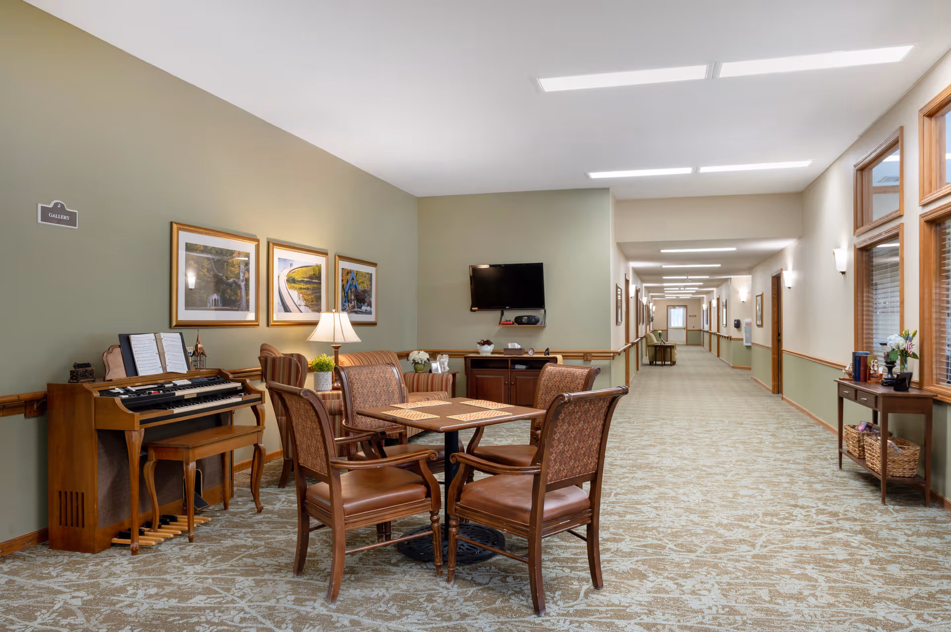 Carpeted senior living hallway with a seating area of table and chairs, an organ, wall art, and a wall-mounted TV.