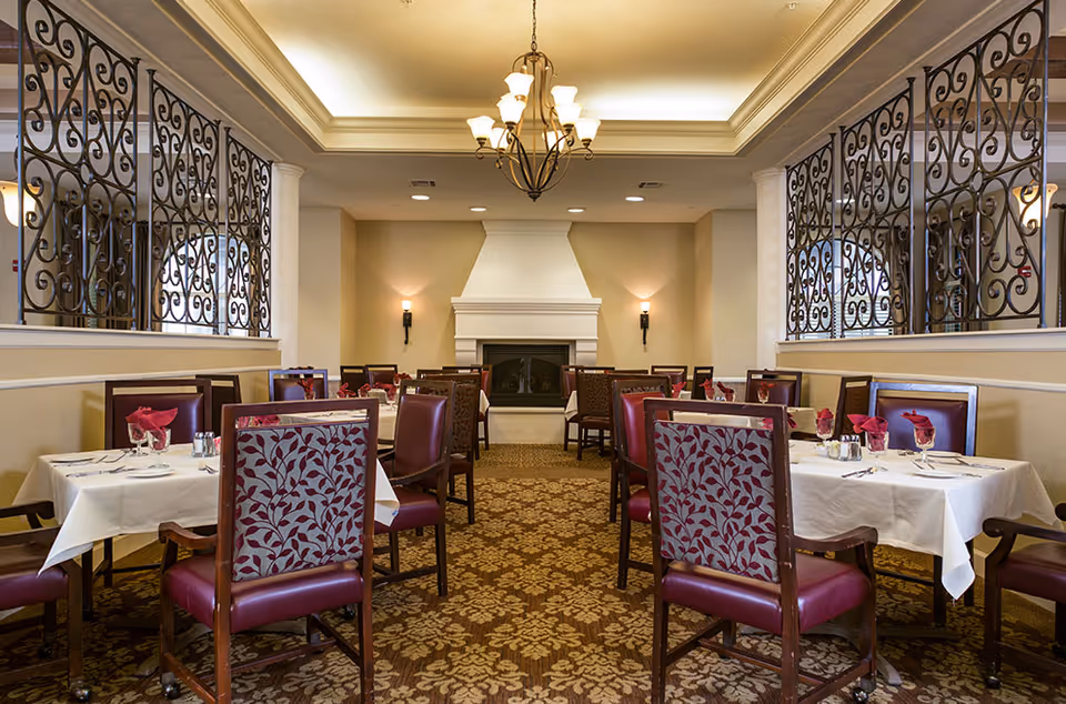Elegant dining room with tables set with white linens and red-upholstered chairs, a chandelier overhead and a fireplace at the far wall.