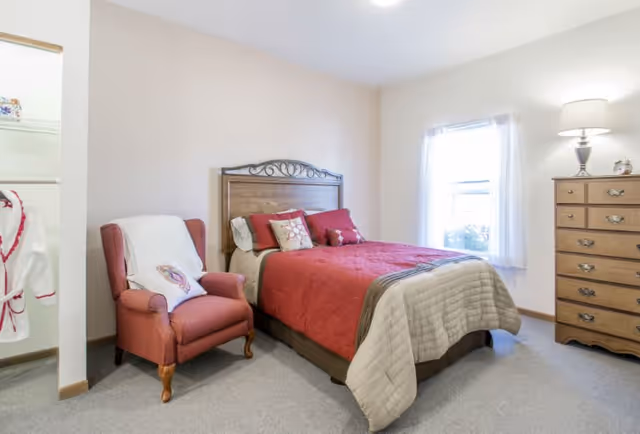 A cozy bedroom with a wooden headboard bed dressed in red and beige bedding, a red upholstered armchair with a white throw blanket, a wooden dresser with a lamp on top, and a window with sheer white curtains letting in natural light.
