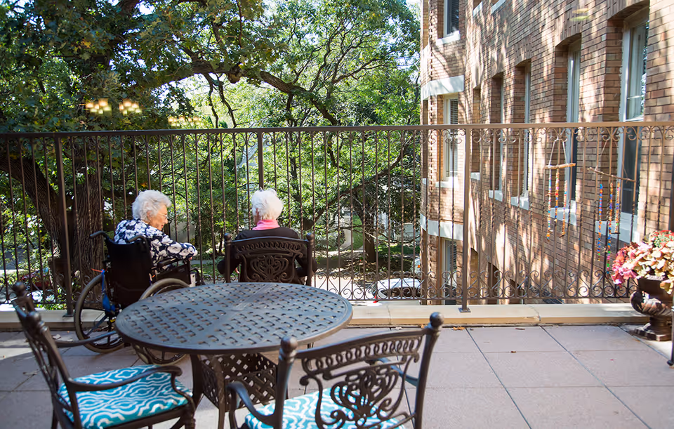 Two elderly women sitting on a balcony with ornate metal railing, one in a wheelchair and the other in a chair, overlooking a green outdoor area with trees. The balcony has a round metal table and chairs with blue patterned cushions, and a brick building is visible on the right side.