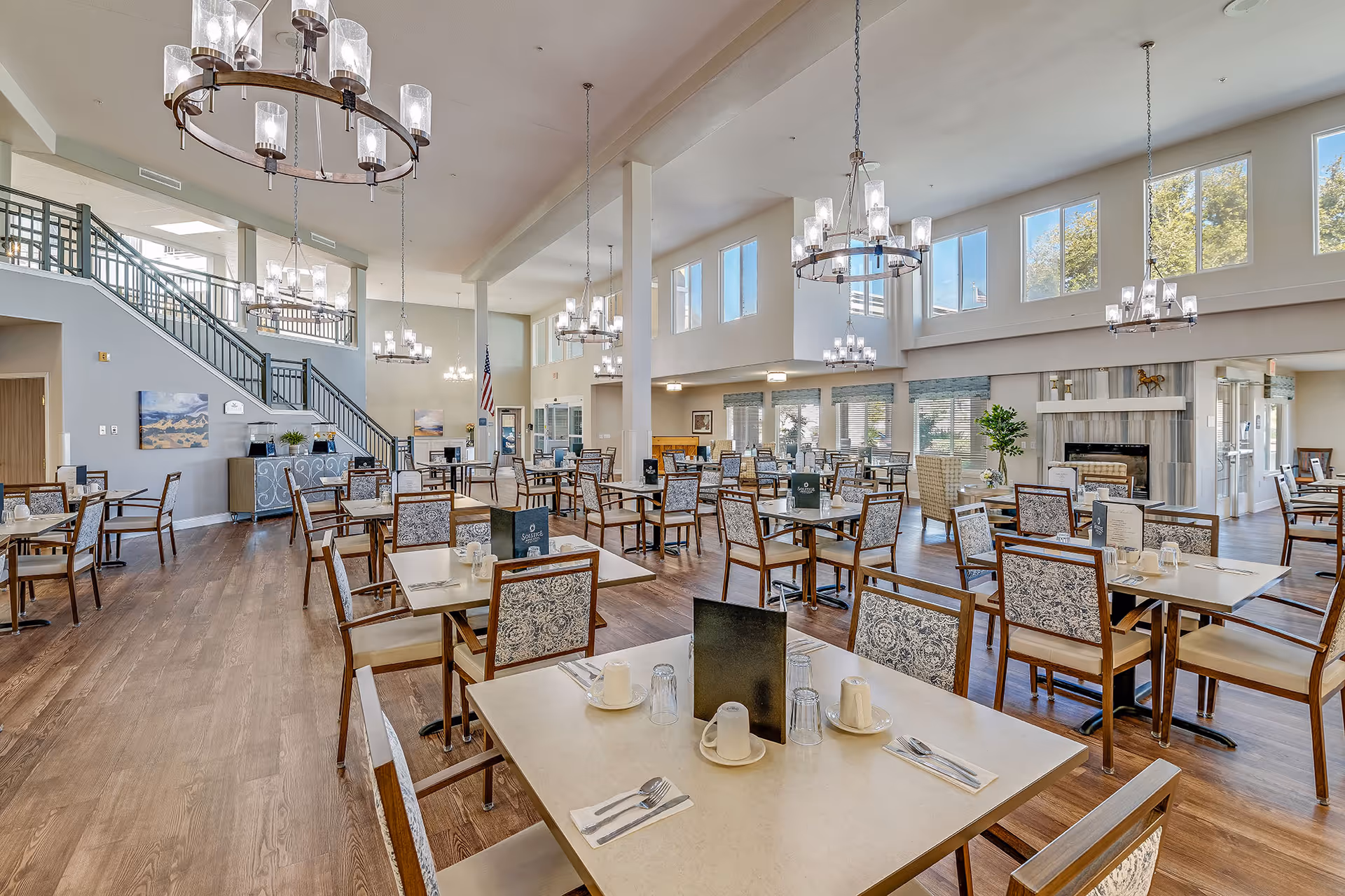 Spacious, well-lit dining room with many set tables, chandeliers, a fireplace and high ceilings in a senior living facility.