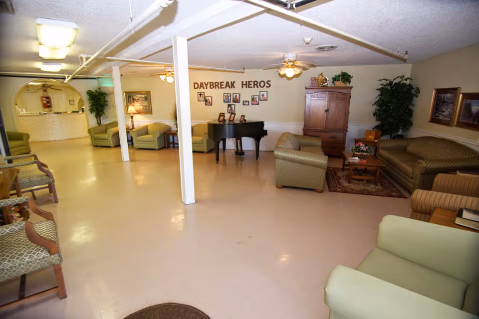 A spacious common area in a senior living facility with multiple armchairs and sofas arranged around a coffee table and a piano. The walls are decorated with framed pictures and the words 'DAYBREAK HEROS'. There are ceiling fans and potted plants adding to the cozy atmosphere.