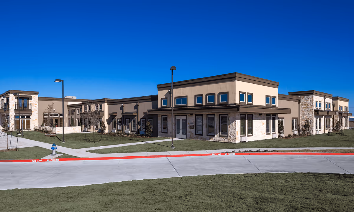 Exterior view of a modern senior living facility building with beige and stone facade, multiple windows, and a clear blue sky. The building is surrounded by green grass, a concrete driveway, and a red-painted fire lane curb.