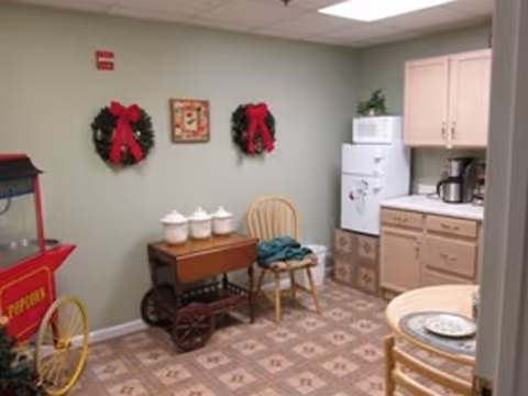 A small kitchen area with beige cabinets, a white refrigerator, microwave, and coffee maker. There is a wooden cart with three white canisters on it, a wooden chair with a green cloth draped over it, and a round table with chairs. The walls are light green with two Christmas wreaths decorated with red bows and a small framed picture between them. A red popcorn machine is partially visible on the left side.