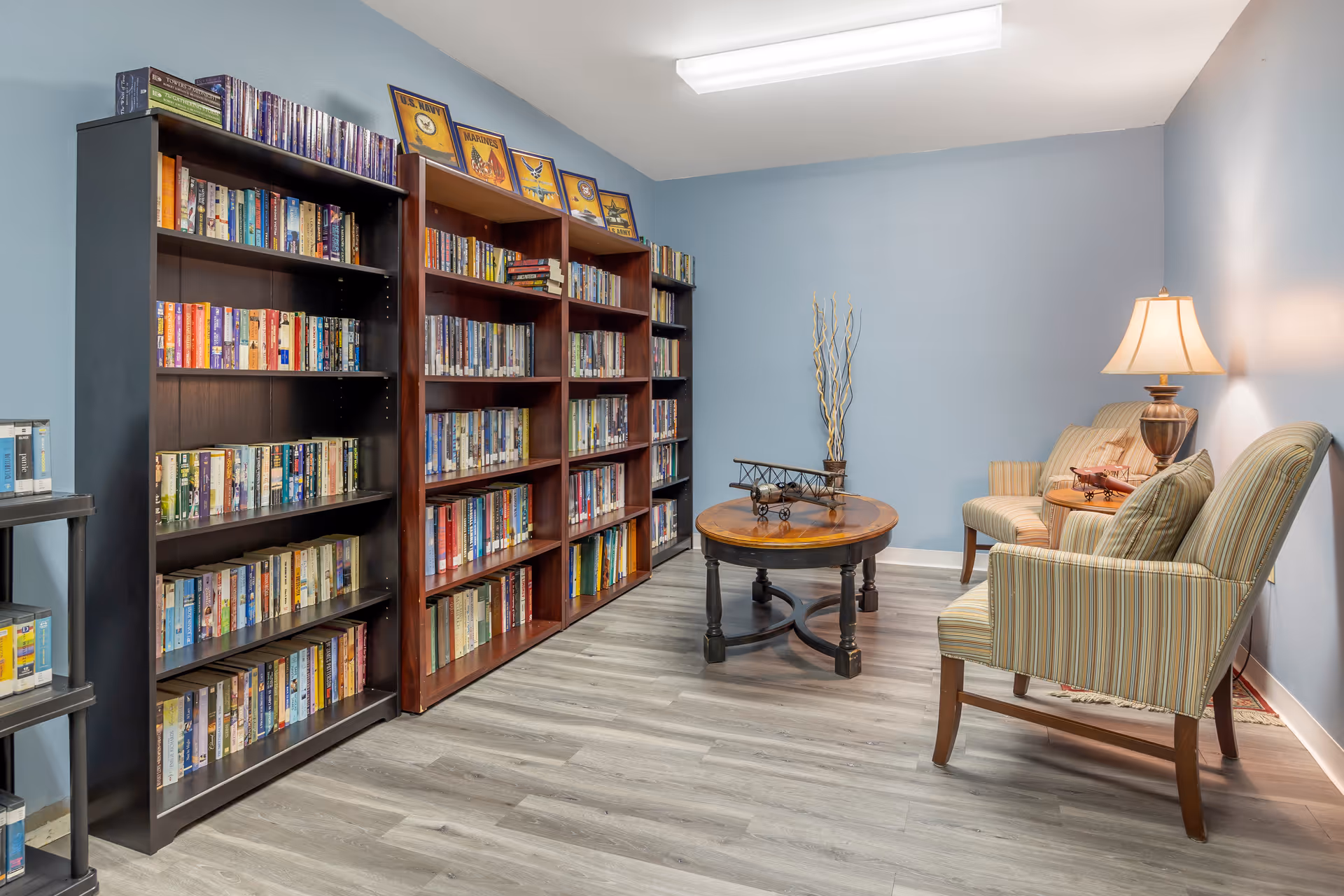A cozy reading room with light blue walls, featuring three large bookshelves filled with books on the left side. On the right side, there are two striped armchairs and a small sofa with a table lamp on a side table. A round wooden coffee table with a decorative model airplane sits in the center of the room. The floor is covered with light wood laminate.